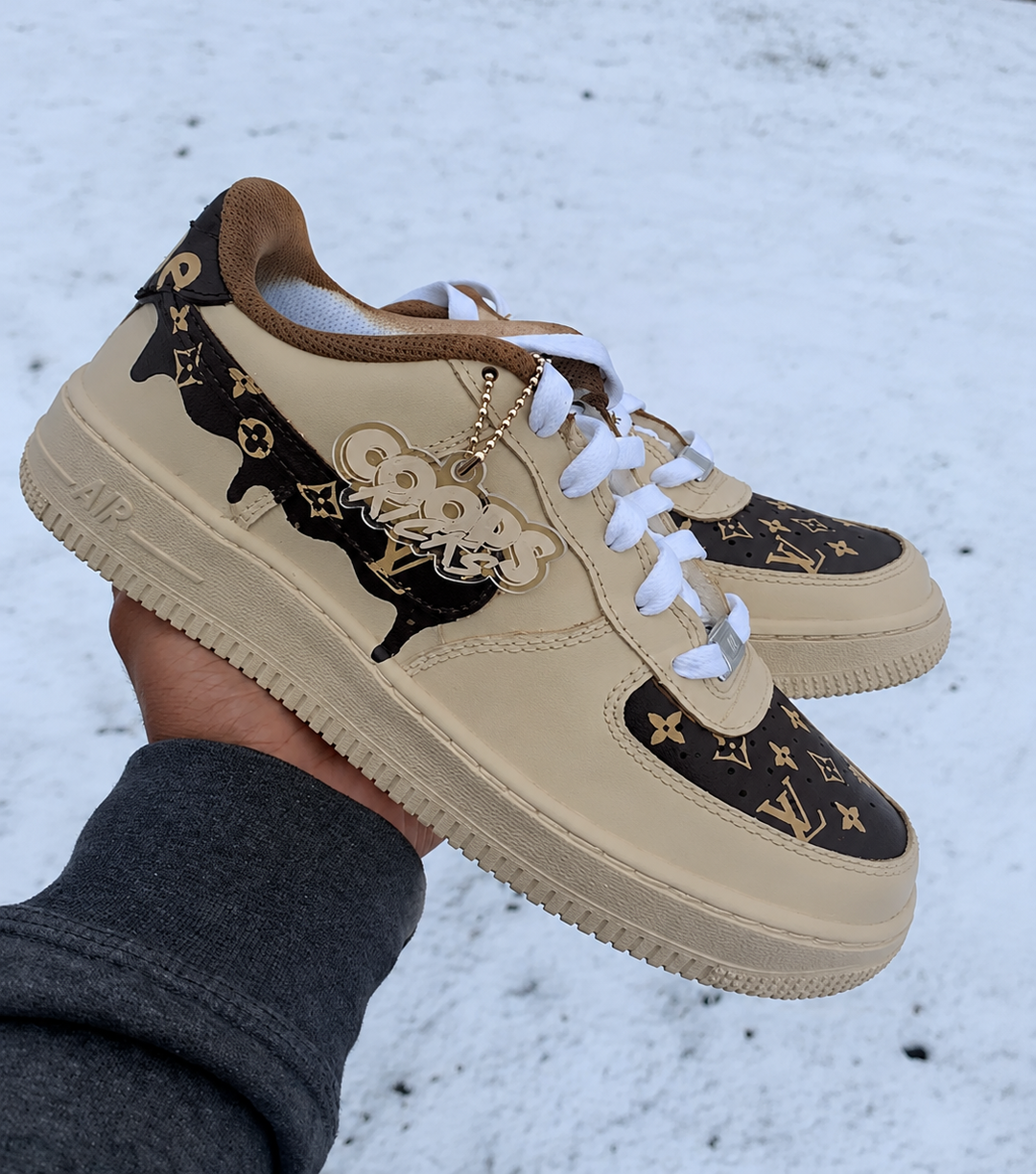 A hand holds a pair of beige and brown monogram sneakers with white laces in an outdoor snowy setting.
